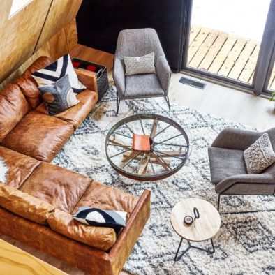 Living room of Aframe house with leather sectional sofa and two lounge chairs from Article on a textured rug and glass coffee table.
