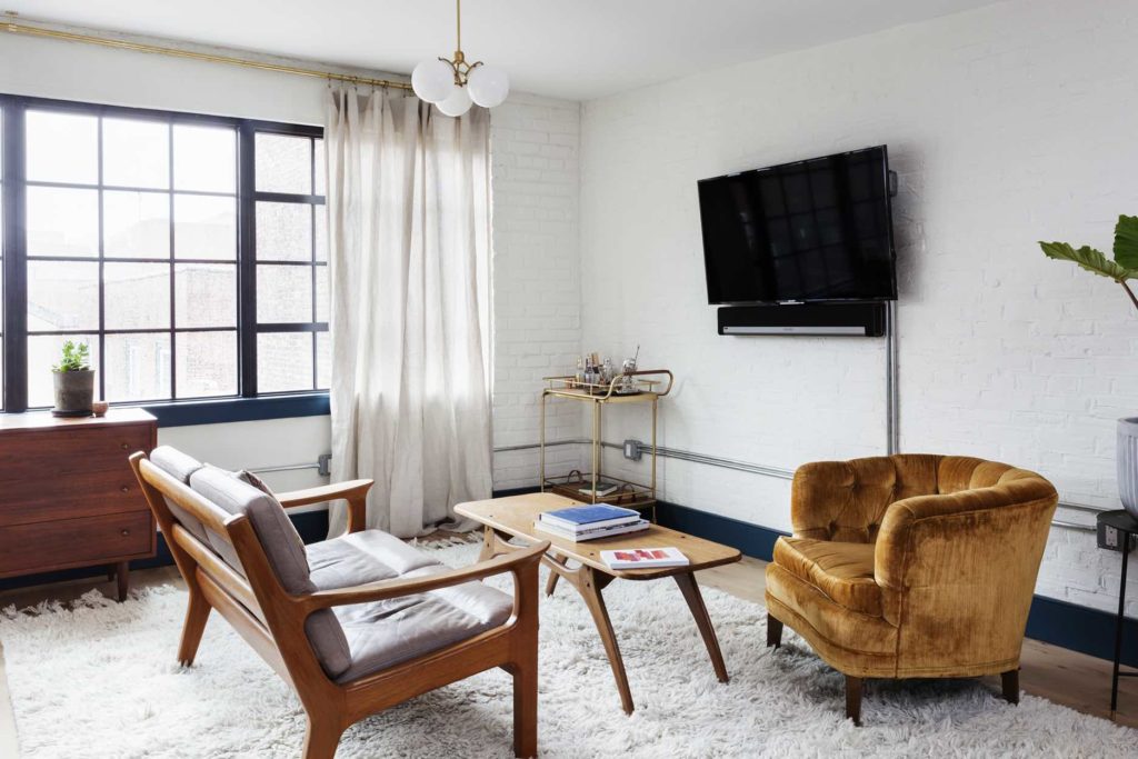 Mid century living room with leather and wood couch, wooden coffee table, plush vintage rug, brass light fixture and yellow fabric chair with factory windows pouring in light behind
