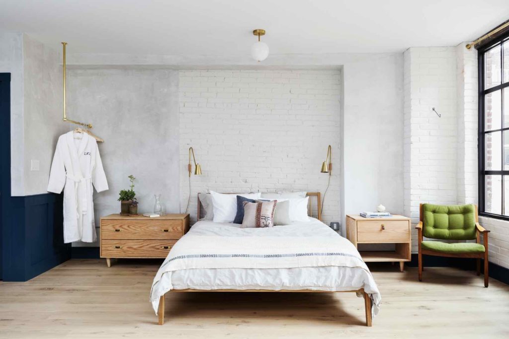 An open loft bedroom in the Will suite at Lokal Hotel Old City with robes hanging on brass hardware, wooden side tables on either side of queen bed and brass bedside light fixtures and green sitting chair