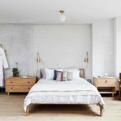 An open loft bedroom in the Will suite at Lokal Hotel Old City with robes hanging on brass hardware, wooden side tables on either side of queen bed and brass bedside light fixtures and green sitting chair