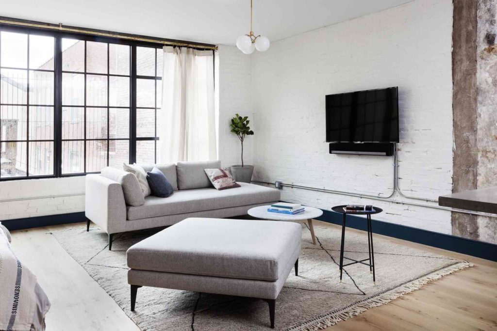 Living area of an apartment suite at Lokal Hotel Old City with couch and ottoman on top of vintage rug with tv on the wall with a sonos speaker and large factory windows flooding the room with light