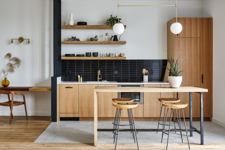 White oak kitchen in Lokal Hotel Fishtown with matte black tile backsplash built in refrigerator white tile floors and an island table that doubles as a dining table with wood and steel stools