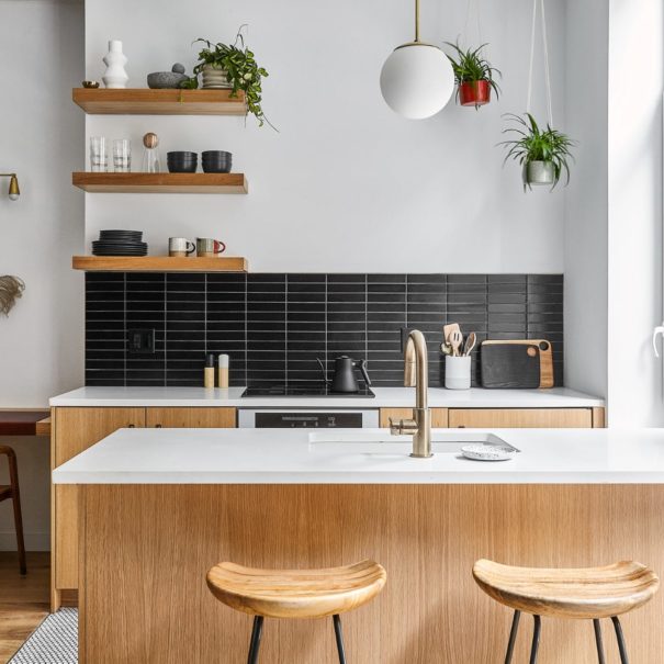 white oak kitchen with island and white quartz countertops floating shelves and brass light fixture with hanging plants in the corner and two stools behind the island and ADA sitting desk to the left