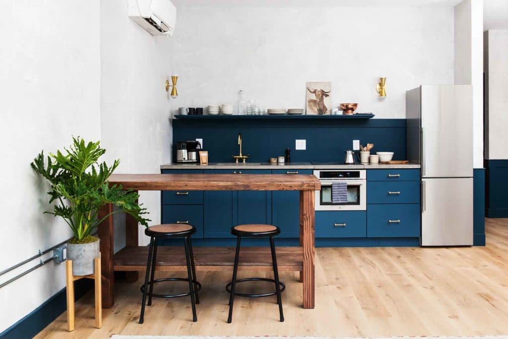 Blue kitchen in apartment hotel room by Lokal Hotel with brass hardware, wide plank white oak floors and custom wooden island with school stools