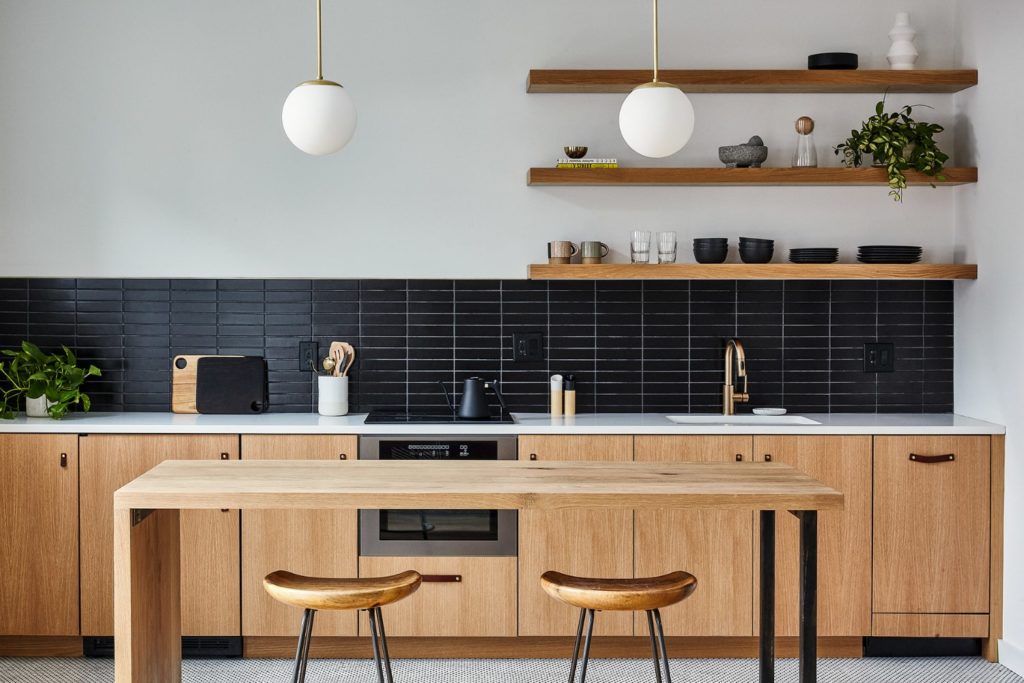 An efficient studio apartment hotel kitchen with white oak built in appliances a black tile backsplash and floating shelves with steel and wood island and stools and brass pendant lights