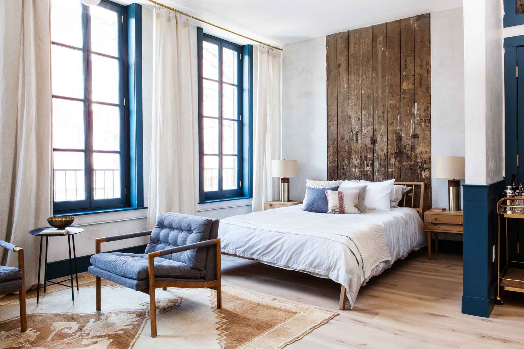 Floor to ceiling historic windows flood this studio apartment hotel room at Lokal Hotel Old City with light with queen bed in front of reclaimed wood accent wall and sitting area with two vintage chairs on a plush area rug